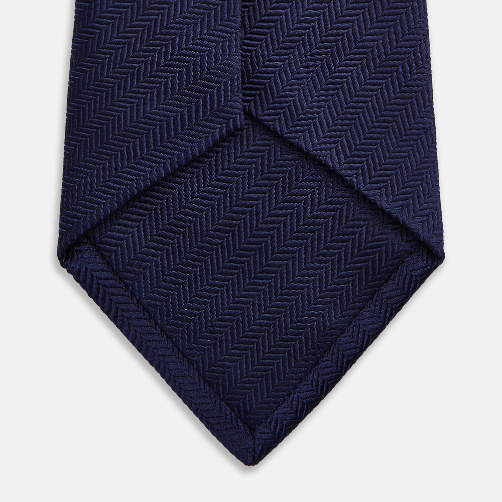 The Turnbull & Asser Slim Navy Herringbone Silk Tie is a refined men’s accessory, elegantly rolled and displayed on a plain white background.