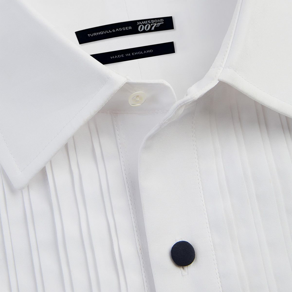 View 8 of product The Casino Royale White Dress Shirt in Cotton by Turnbull & Asser UK is a long-sleeve white dress shirt, crafted from crisp white cotton poplin with a pleated bib front, black buttons, and a classic collar, shown laid flat on a white background.