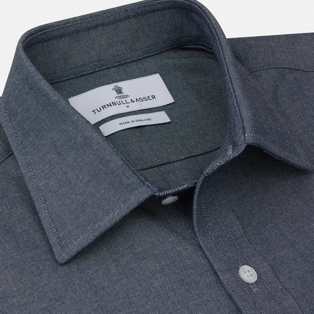 The Turnbull & Asser Plain Blue Oxford Weekend Fit Shirt features a Derby collar, 1-button cuffs, and a smart-casual look—perfect for weekends. Displayed flat on a white background.