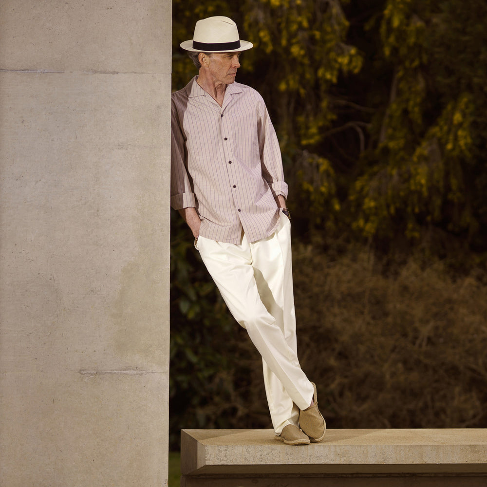 An older man in a light hat, Turnbull & Asser Beige Stripe Cotton Holiday Fit Shirt with Revere Collar & Single Button Cuff, and white pants leans casually against a tall concrete pillar outdoors, greenery blurred behind him.