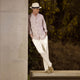 An older man in a light hat, Turnbull & Asser Beige Stripe Cotton Holiday Fit Shirt with Revere Collar & Single Button Cuff, and white pants leans casually against a tall concrete pillar outdoors, greenery blurred behind him.