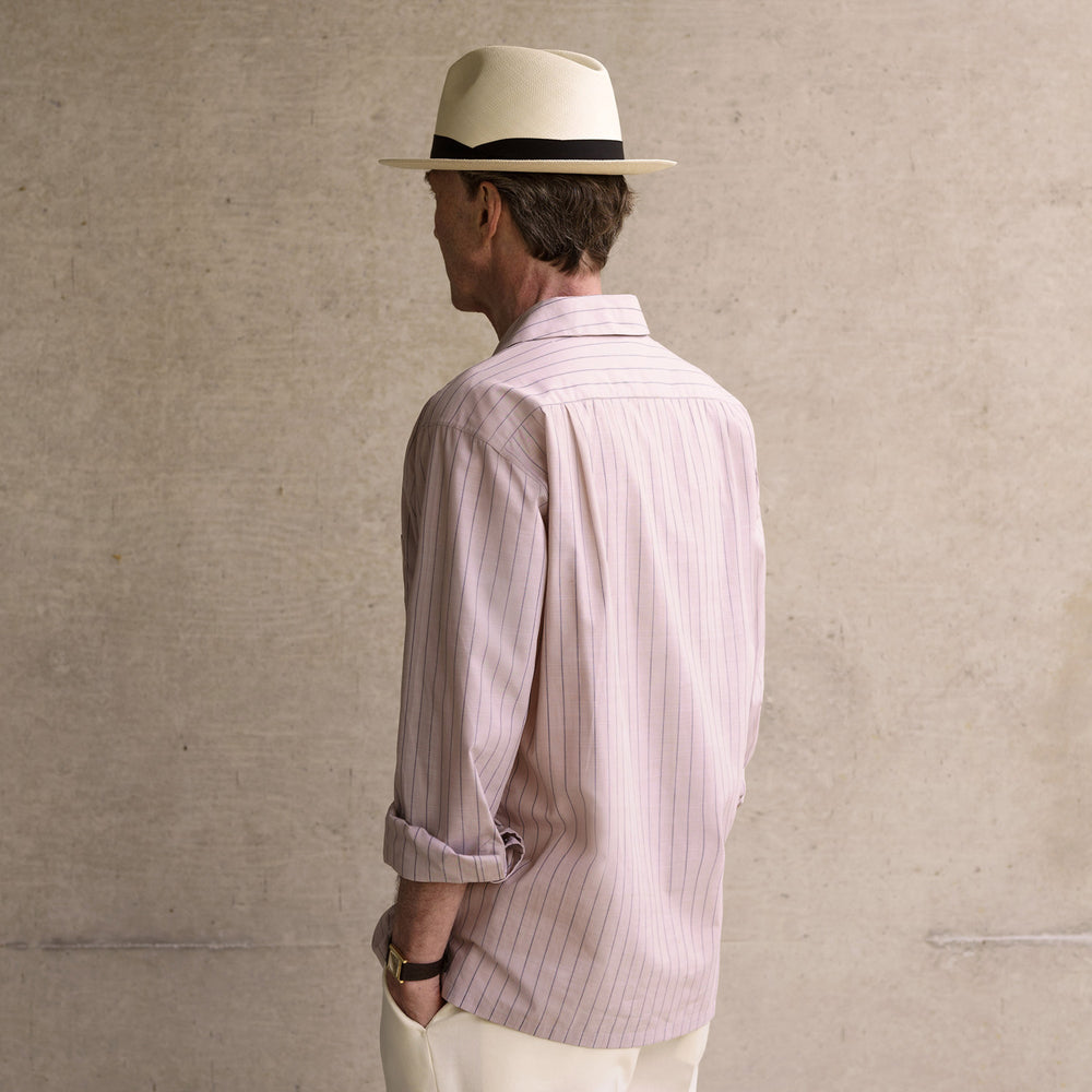 An older man in a light hat, Turnbull & Asser Beige Stripe Cotton Holiday Fit Shirt with Revere Collar & Single Button Cuff, and white pants leans casually against a tall concrete pillar outdoors, greenery blurred behind him.