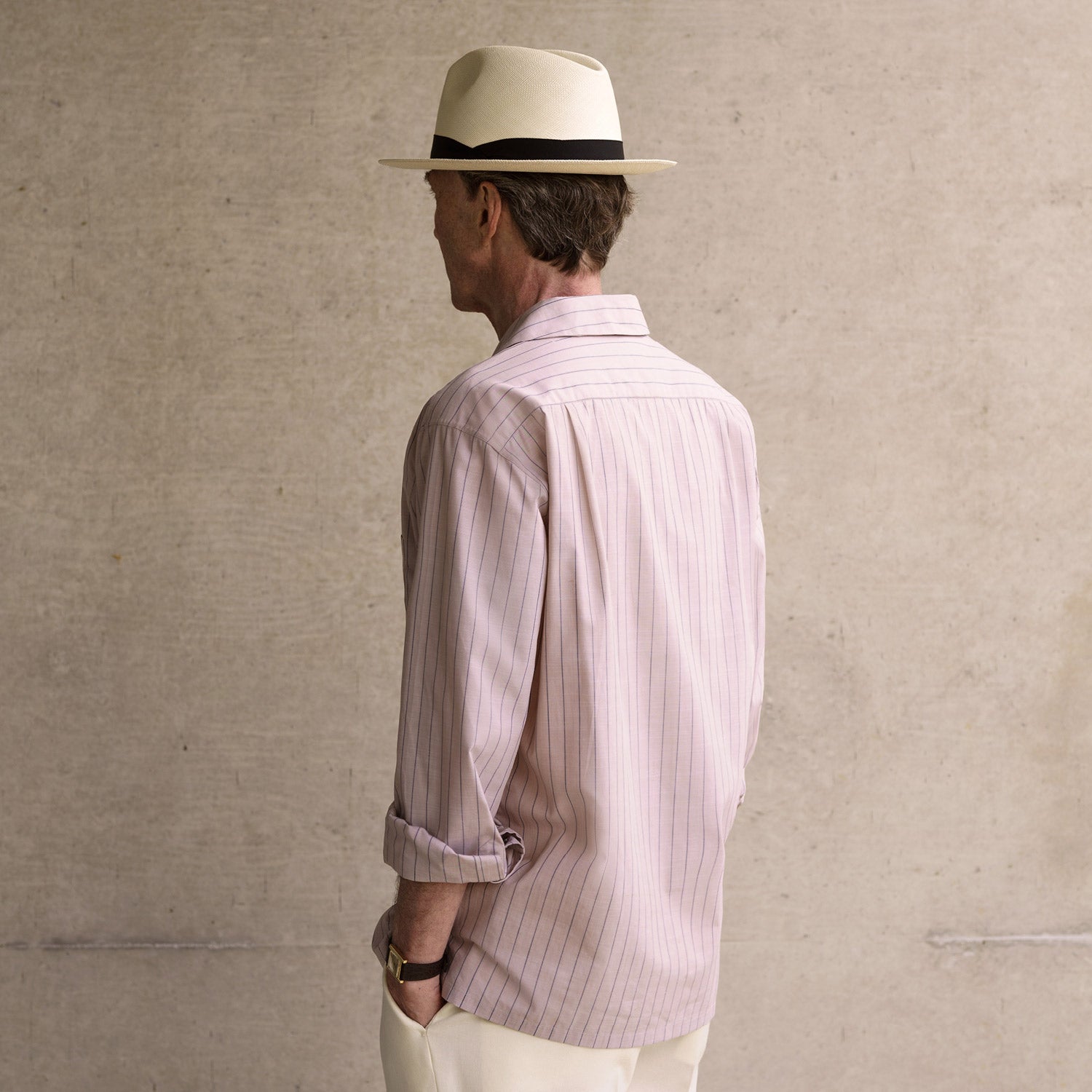 View 3 of product An older man in a light hat, Turnbull & Asser Beige Stripe Cotton Holiday Fit Shirt with Revere Collar & Single Button Cuff, and white pants leans casually against a tall concrete pillar outdoors, greenery blurred behind him.