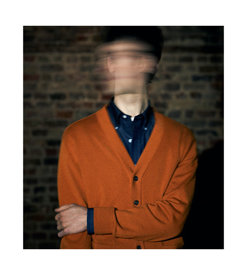 A person in an orange cardigan and blue shirt stands against a brick wall, with their face blurred by motion, creating a sense of dynamic movement or obscured identity.