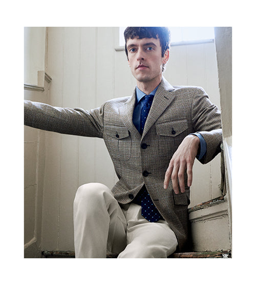 A man with short dark hair sits on stairs, wearing a gray checkered blazer, blue dress shirt, blue polka dot tie, and light-colored pants. He looks forward with a neutral expression, arm resting on the stair rail.
