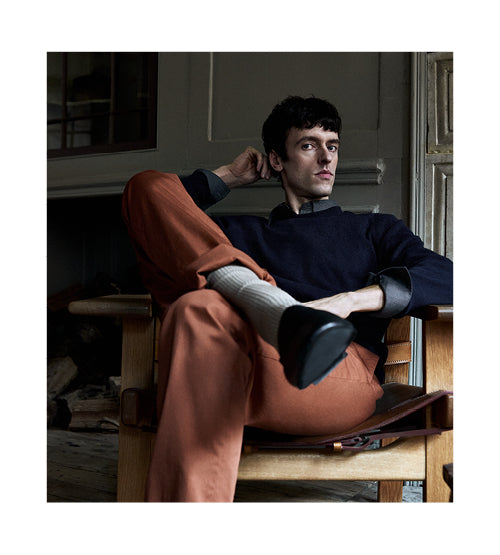 A person with short dark hair sits casually in a wooden chair indoors, wearing an indigo sweater, orange pants, light socks, and black shoes, with one arm resting behind their head and legs crossed.
