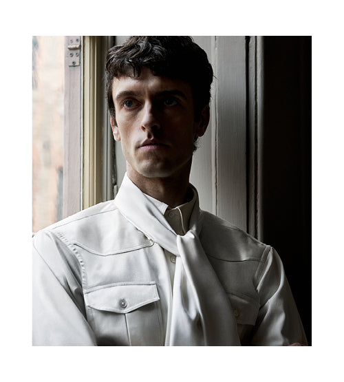 A man with short dark hair wears a white Western-style shirt and a loosely tied white scarf, standing by a window with light softly illuminating his face.