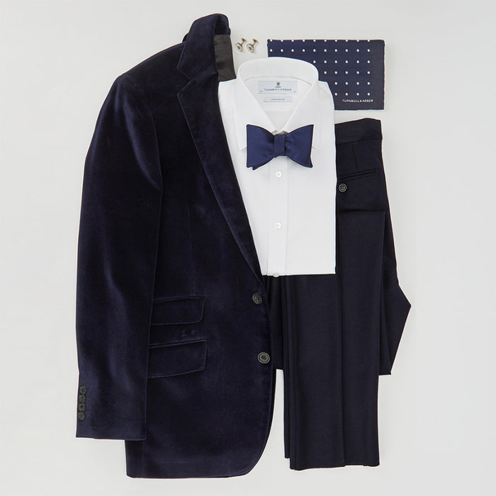 A neatly arranged navy blue velvet suit with matching trousers, a white dress shirt, a navy blue bow tie, and a navy pocket square with white dots, all laid out on a flat white background.