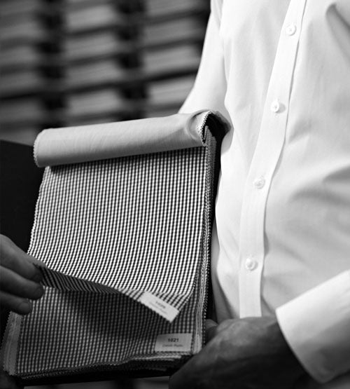 A person in a white shirt holds a fabric swatch book open, displaying a checkered pattern fabric sample while another hand lifts a swatch. The background is blurred with stacked shelves. Black and white image.