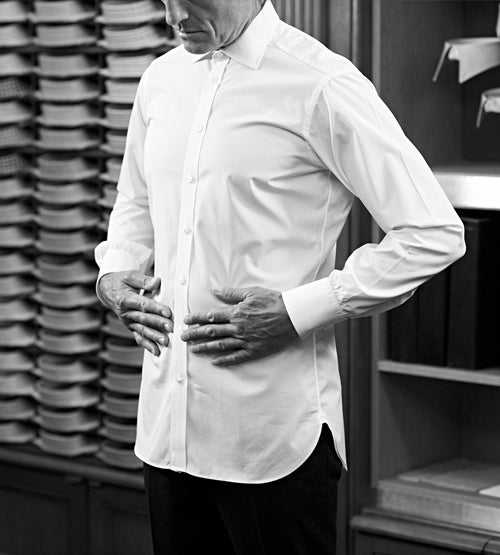A man wearing a white dress shirt stands in a clothing store, touching his stomach with both hands. Shelves of folded shirts are visible in the background. The image is in black and white.
