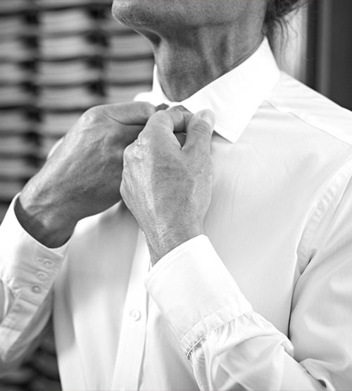 A person in a white dress shirt adjusts their collar, preparing to button or fasten it. The image is in black and white and focuses on their hands and shirt, with their face partially out of frame.