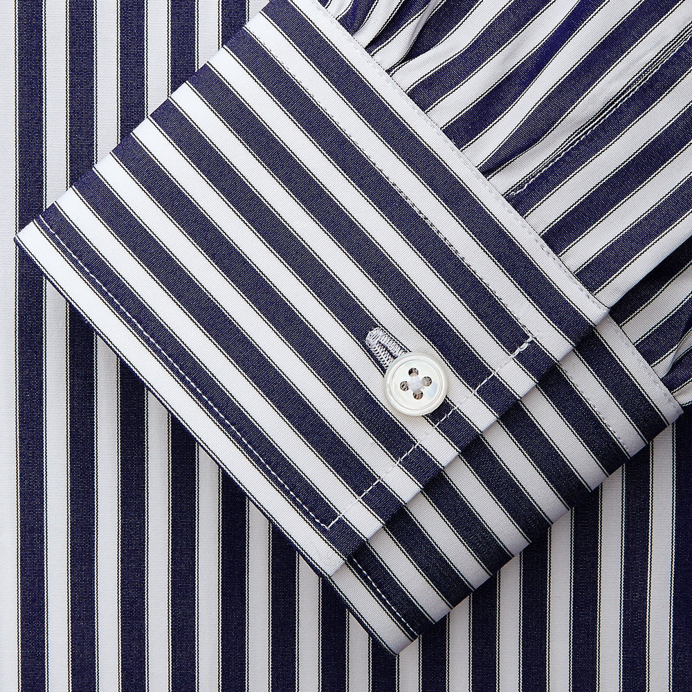 A person in a Turnbull & Asser Navy & White Stripe Weekend Fit Shirt with Derby Collar and Single Button Cuffs, paired with a paisley tie and dark pants, stands indoors leaning against a cream paneled wall.