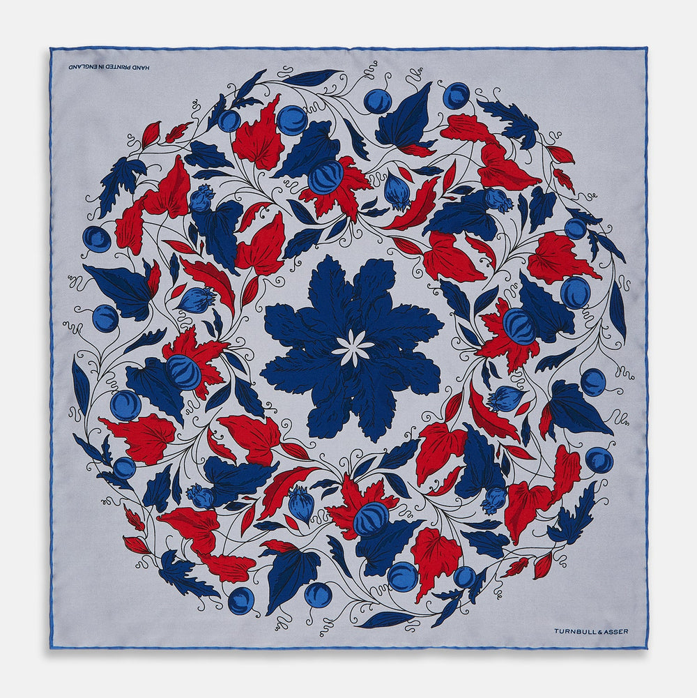 The Blue and Red Foliage Pattern Silk Pocket Square by Turnbull & Asser features intricate symmetrical leaves and flowers in red, blue, and navy on a light grey silk background, creating an elegant circular motif.