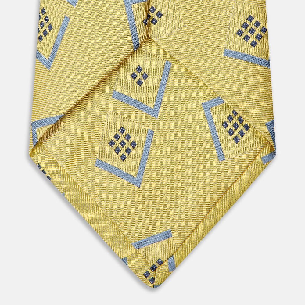 The Turnbull & Asser Yellow Graphic Deco Silk Tie features a subtle pattern of light blue geometric shapes and small black dots, elegantly rolled against a white background.