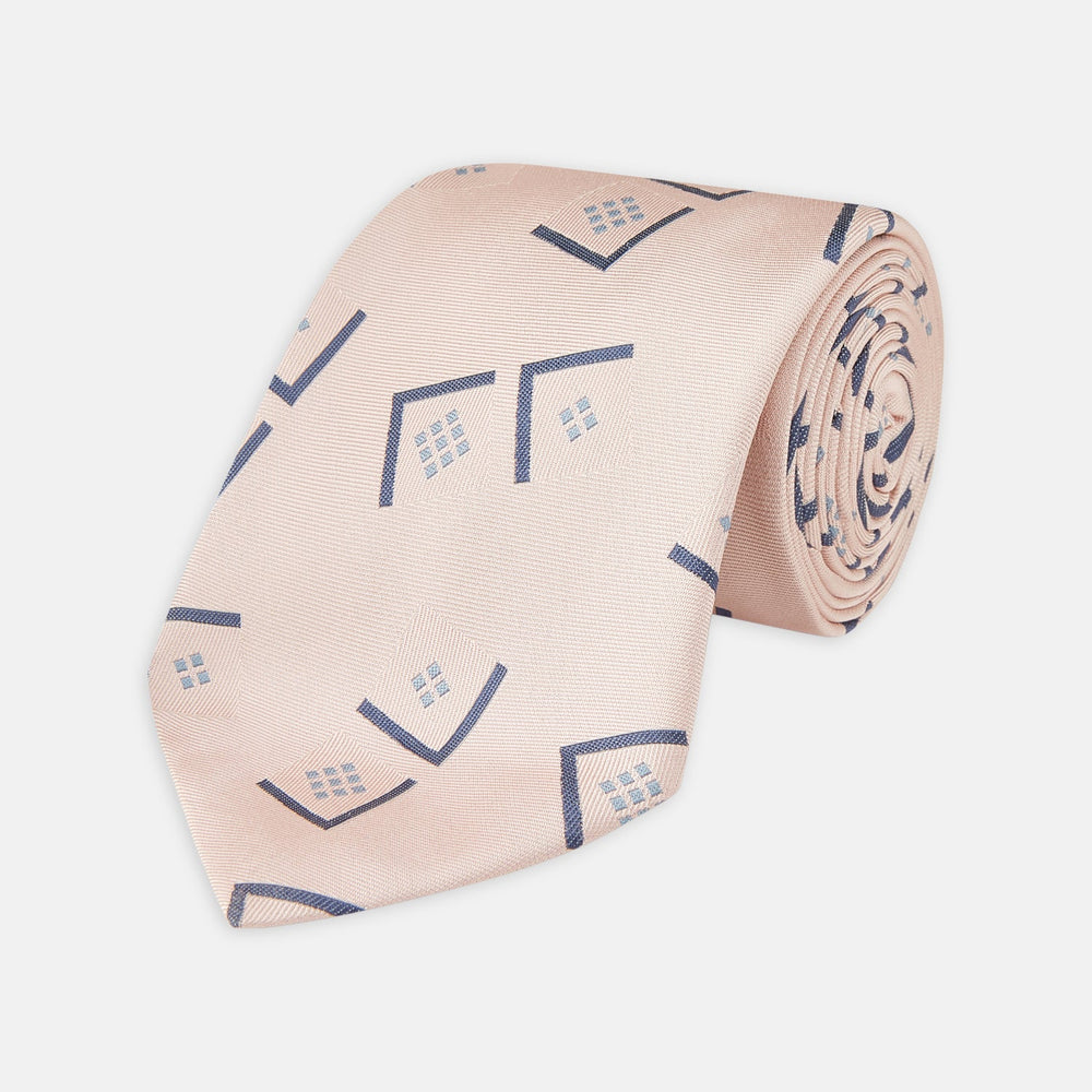 The Turnbull & Asser Pink Graphic Deco Silk Tie features blue geometric and square patterns on pale pink silk, shown rolled with the pointed end facing forward.