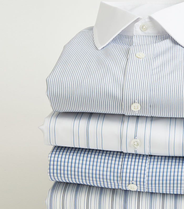 A neatly folded stack of four mens dress shirts in blue and white patterns, including stripes and checks, with the top shirt featuring a white collar.