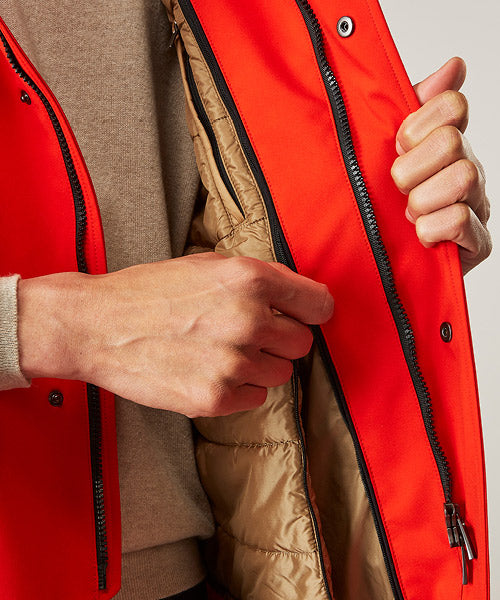 A person unzips a bright orange jacket, revealing a tan quilted inner lining. Only their hands and part of their beige sweater sleeve are visible.