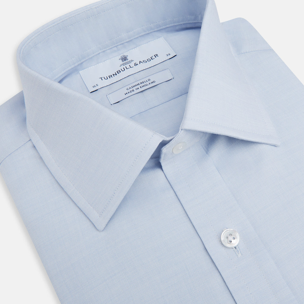 The Turnbull & Asser Blue Cashmerello Mayfair Shirt is a light blue cotton-cashmere blend with long sleeves, pointed collar, white buttons, and buttoned cuffs, shown on a plain white background.