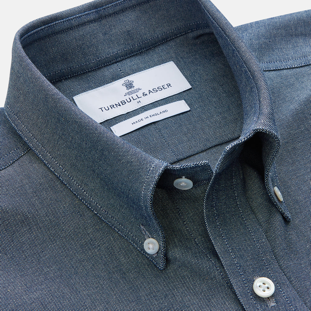 The Turnbull & Asser Blue Chambray Weekend Fit Nevis Cotton Shirt features a Dorset collar, 1-button cuffs, and smart-casual tailoring, shown laid flat on a white background.