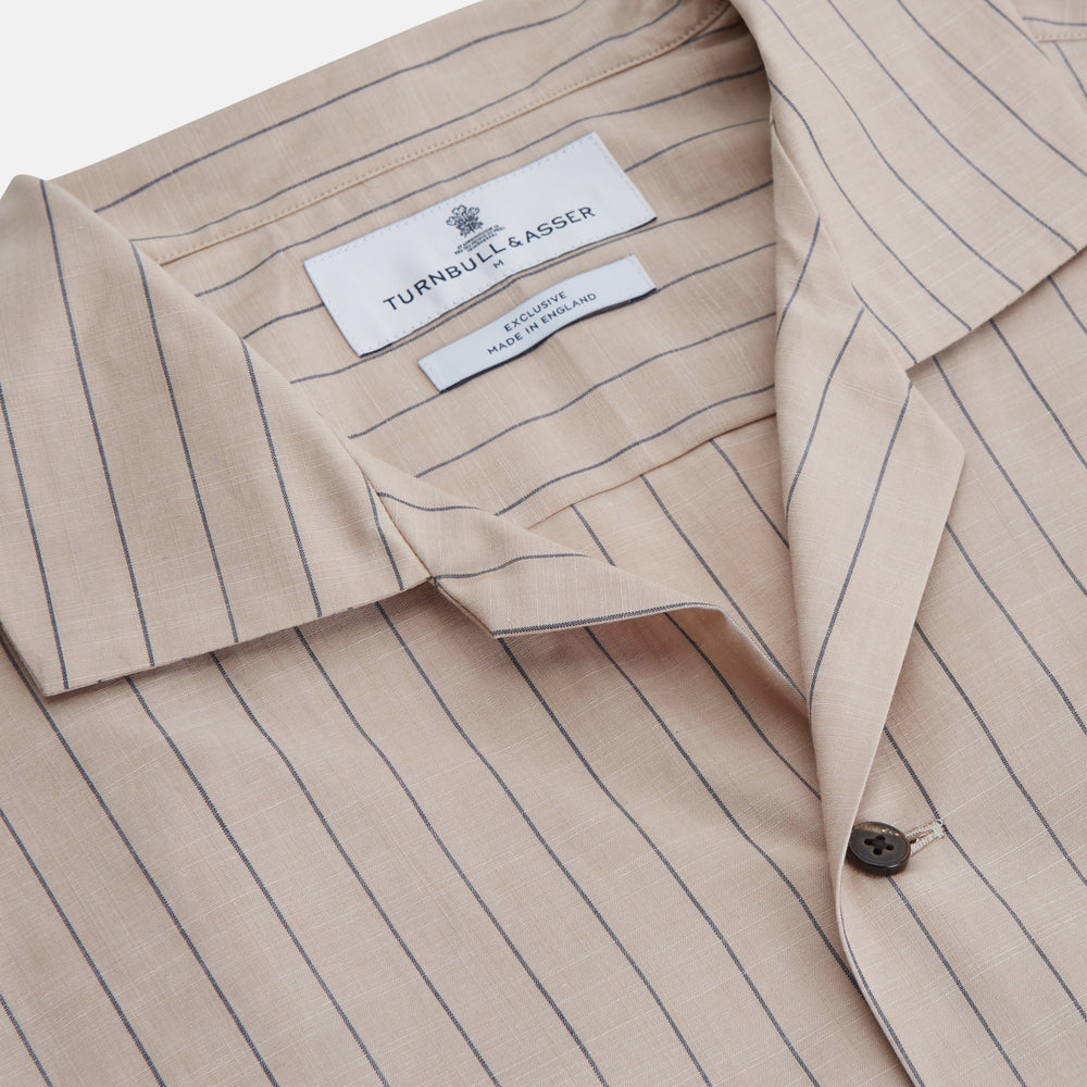 An older man in a light hat, Turnbull & Asser Beige Stripe Cotton Holiday Fit Shirt with Revere Collar & Single Button Cuff, and white pants leans casually against a tall concrete pillar outdoors, greenery blurred behind him.