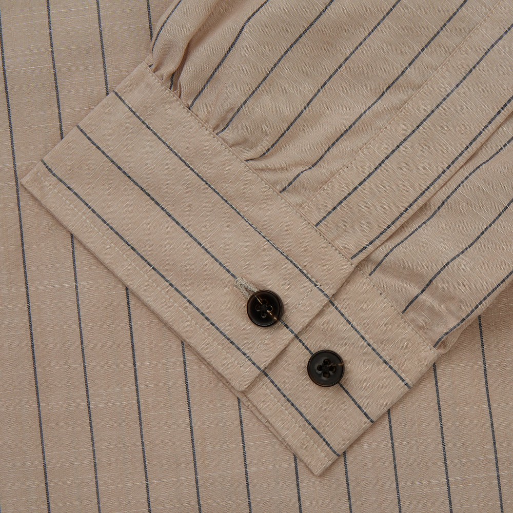 An older man in a light hat, Turnbull & Asser Beige Stripe Cotton Holiday Fit Shirt with Revere Collar & Single Button Cuff, and white pants leans casually against a tall concrete pillar outdoors, greenery blurred behind him.