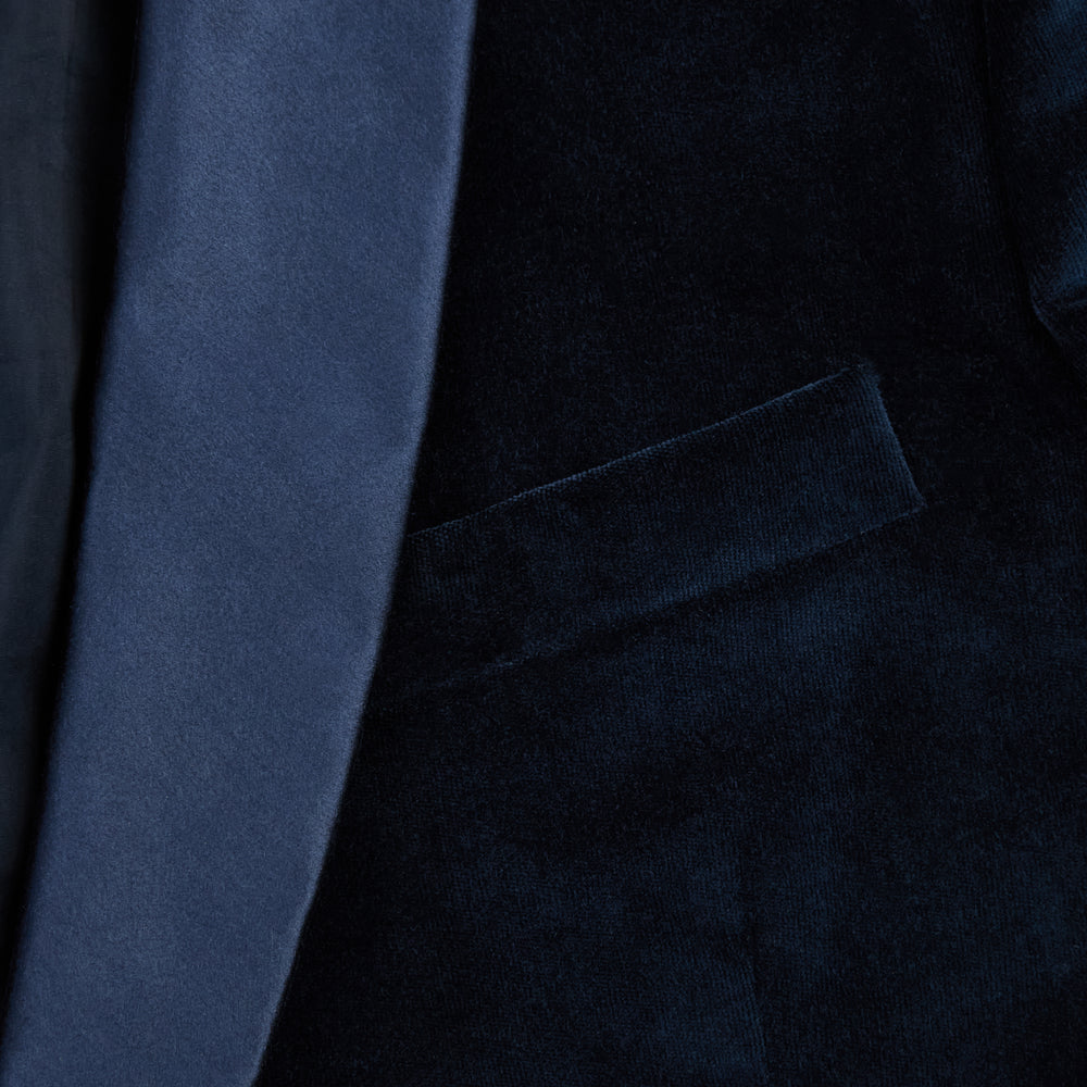 Turnbull & Asser UK’s Casino Royale Midnight Blue Tuxedo Jacket in Cotton Velvet, featuring a single button closure and a luxurious silk-satin shawl lapel, is showcased against a plain white background.