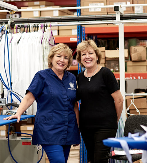 Two women smiling and standing in a workspace with clothing racks, boxes, and equipment in the background. One woman wears a blue uniform; the other wears a black top. The setting appears to be a garment or laundry facility.