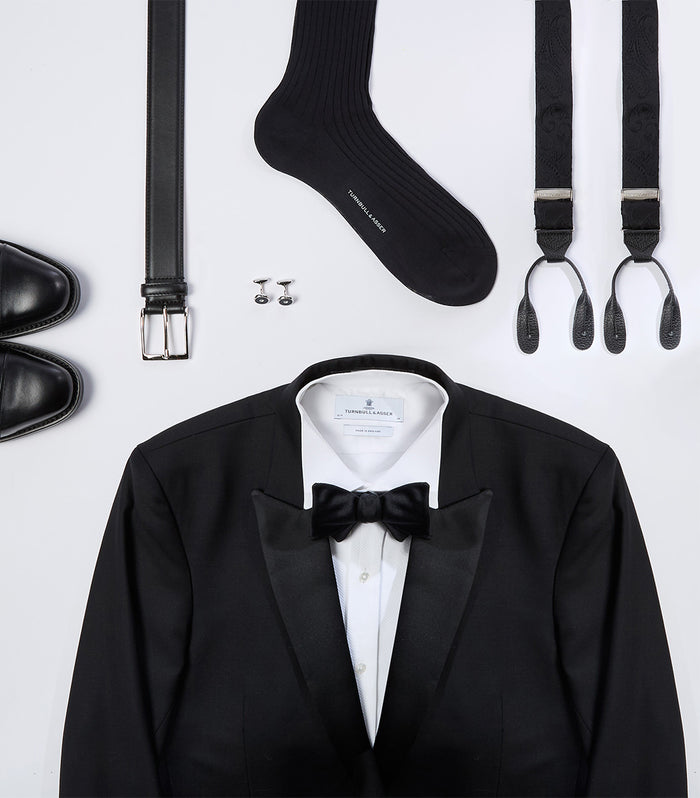 Flat lay of formal wear items, including a black tuxedo jacket with a bow tie and white shirt, black dress shoes, a belt, black socks, cufflinks, and suspenders, all arranged neatly on a white background.