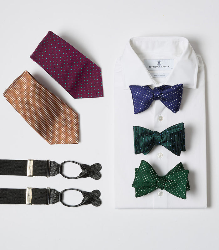 A white dress shirt with three polka dot bow ties in blue, green, and teal is displayed alongside two polka dot neckties in burgundy and orange, and a pair of black suspenders.