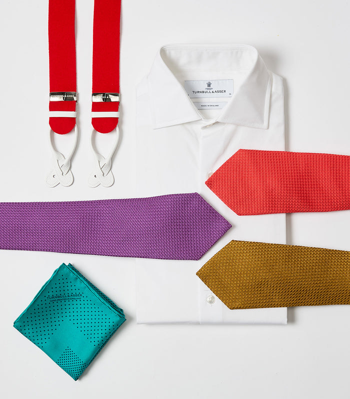 A white dress shirt is neatly folded, surrounded by red suspenders, a purple tie, an orange tie, a mustard yellow tie, and a teal pocket square on a white background.