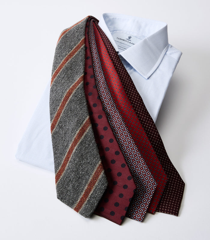 A light blue dress shirt with five neckties draped over it, featuring various patterns: gray with red and tan stripes, maroon with small blue dots, maroon with red dots, and two with geometric designs.