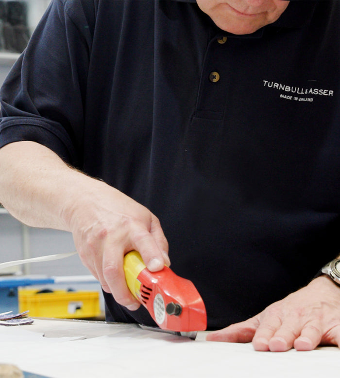 A person in a navy shirt uses a red and yellow utility knife to cut white material on a table, holding the material steady with their other hand.