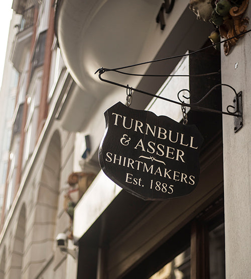 A black hanging sign reads Turnbull & Asser Shirtmakers Est. 1885 outside a building on a city street, with architectural details visible in the background.