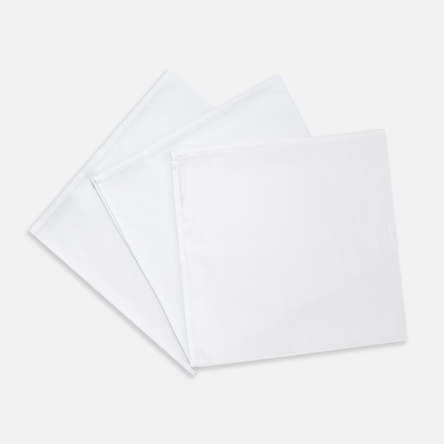 View 1 of product Three Turnbull & Asser Plain White Cotton Handkerchiefs from the 3-pack are neatly folded and overlapping on a light gray background.
