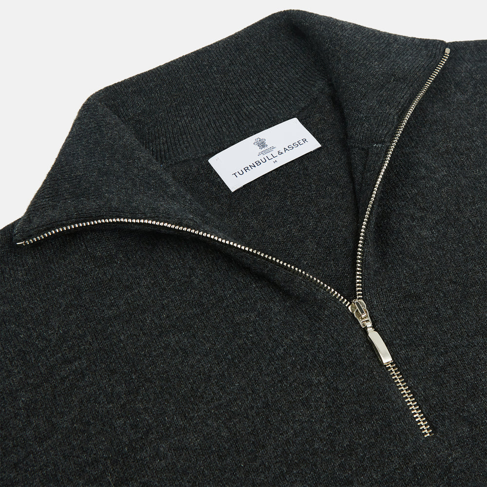 The Turnbull & Asser Charcoal Half-Zip Cashmere Jumper features long sleeves, a high collar, and a zip closure from the collar down the chest. Displayed flat with sleeves extended, it is crafted in dark charcoal grey cashmere.