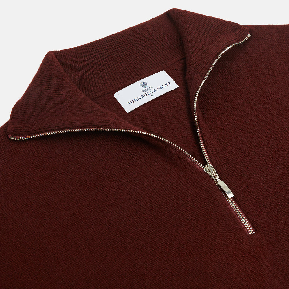 The Turnbull & Asser Port Cashmere Half-Zip Jumper is a dark maroon, long-sleeve style with a high collar, silver half-zip, and ribbed cuffs and hem, shown laid flat on a white background—ideal for layering.