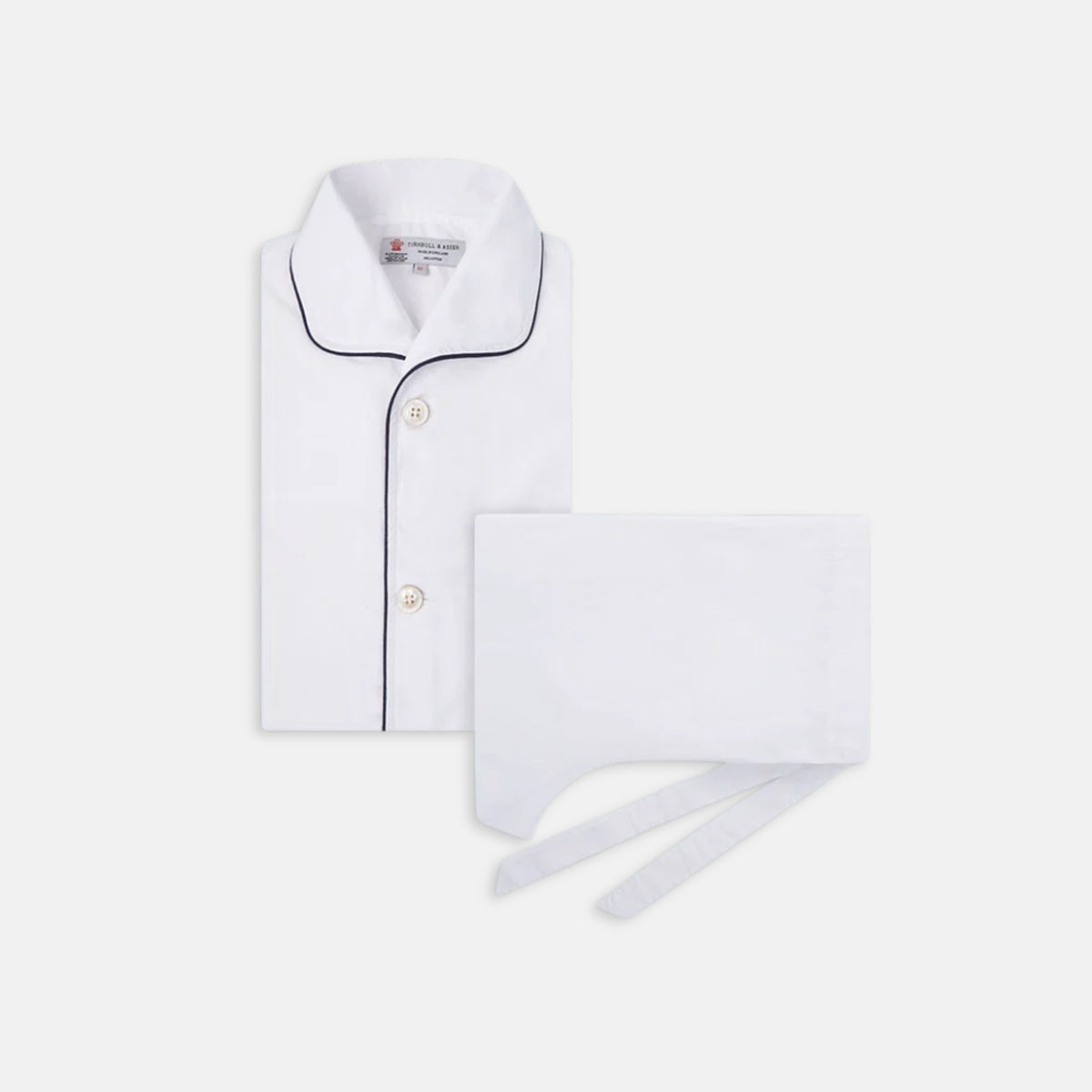 View 1 of product The Turnbull & Asser White Piped Cotton Pyjama Set, made from Egyptian cotton and featuring black piping and two buttons, is shown folded with a matching white tie-strap face mask on a light gray background.