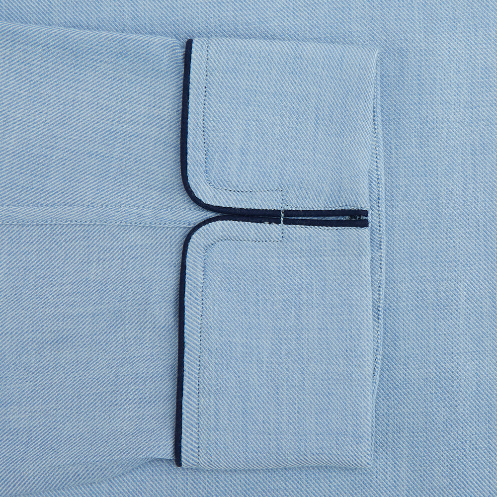 The Blue Piped Cotton-Cashmere Pyjama Set by Turnbull & Asser features a light blue long-sleeved, button-up top with a chest pocket and dark piping, plus matching side-tie pants—luxurious comfort displayed on a white background.
