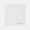 View 3 of product The White Piped Silk Pocket Square by Turnbull & Asser is shown folded at one corner with the brand's logo in black on the lower right, set against a light blue background.