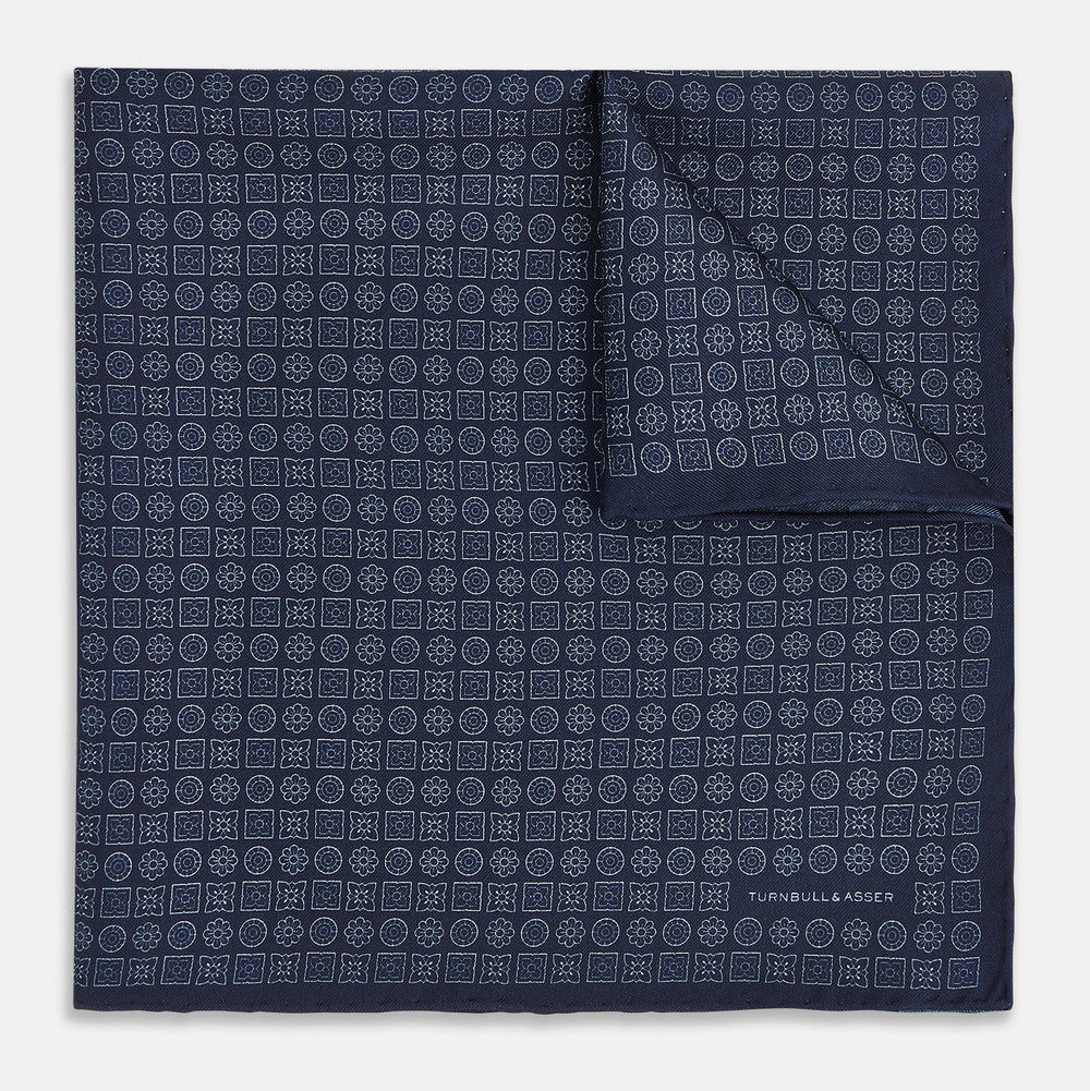 A Floral Navy Pocket Square in Silk by Turnbull & Asser UK, featuring a repeating geometric pattern with the brand’s name printed in white in the corner—perfect for adding a touch of sophistication to formal tailoring. The top right edge is folded over to reveal the reverse side.