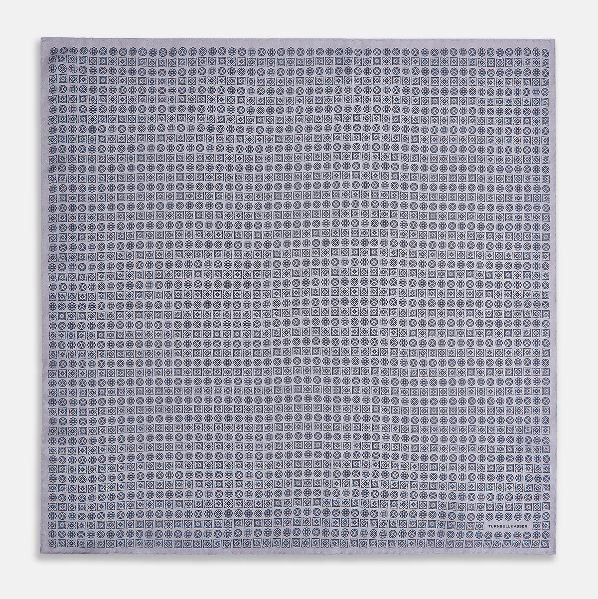 View 4 of product A lilac silk pocket square featuring a repeating geometric rosette pattern. One corner is folded over to show the reverse side, and "Turnbull & Asser UK" is printed in the bottom right corner.