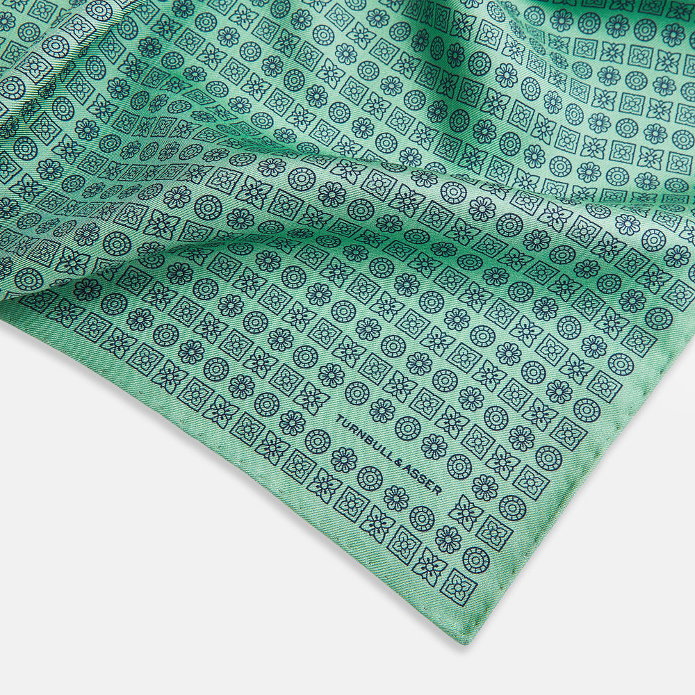 Floral Green Pocket Square in Silk by Turnbull & Asser UK, showcasing navy-blue floral geometric motifs on a bright green background. One corner is neatly folded to display both sides of the silk fabric, with "Turnbull & Asser UK" printed in the lower right corner.