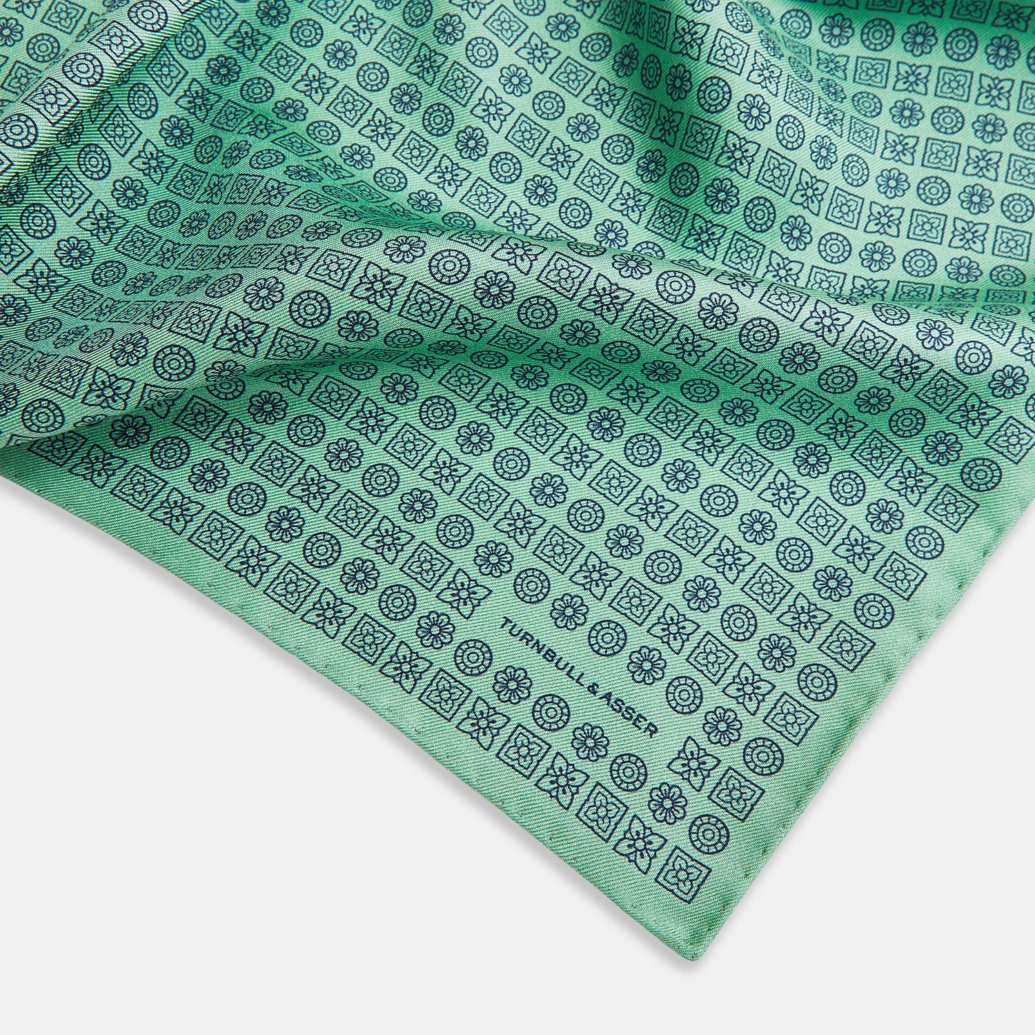 View 2 of product Floral Green Pocket Square in Silk by Turnbull & Asser UK, showcasing navy-blue floral geometric motifs on a bright green background. One corner is neatly folded to display both sides of the silk fabric, with "Turnbull & Asser UK" printed in the lower right corner.