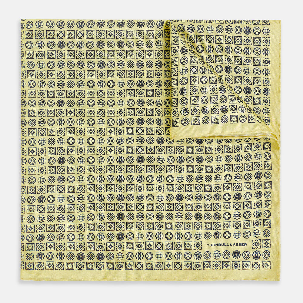 A Floral Yellow Pocket Square in Silk by Turnbull & Asser UK, showcasing a repeating black geometric pattern with subtle floral motifs and the brand logo in one corner. One corner of the fabric is folded over to display the reverse side.