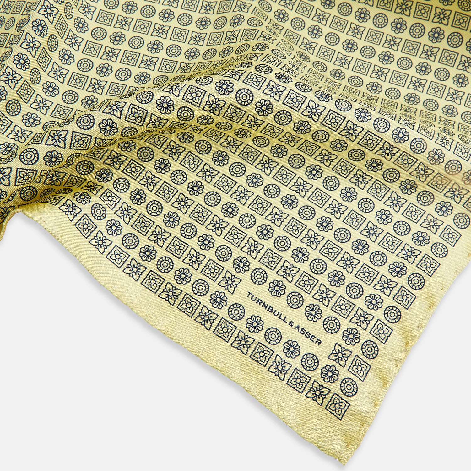 View 2 of product A Floral Yellow Pocket Square in Silk by Turnbull & Asser UK, showcasing a repeating black geometric pattern with subtle floral motifs and the brand logo in one corner. One corner of the fabric is folded over to display the reverse side.
