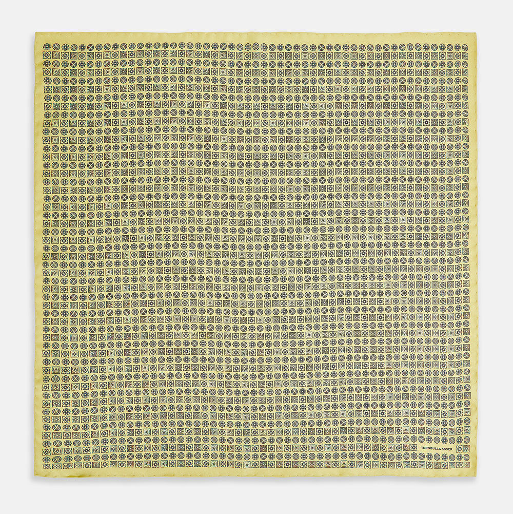 A Floral Yellow Pocket Square in Silk by Turnbull & Asser UK, showcasing a repeating black geometric pattern with subtle floral motifs and the brand logo in one corner. One corner of the fabric is folded over to display the reverse side.