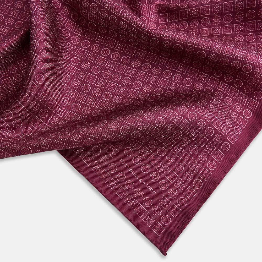 A folded Floral Burgundy Pocket Square in Silk by Turnbull & Asser UK, adorned with pink floral geometric motifs and featuring the Turnbull & Asser logo in the bottom right corner.