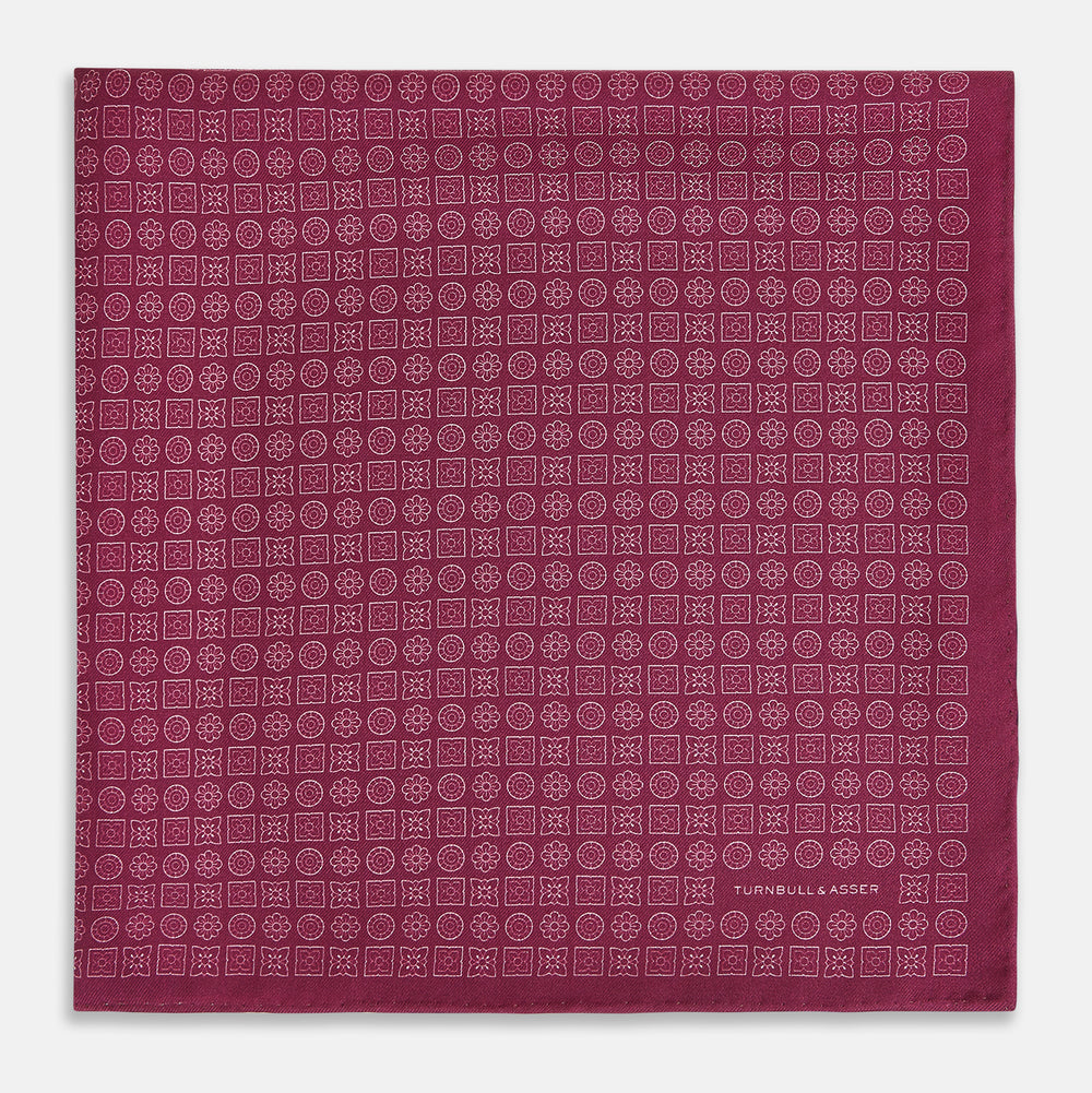 A folded Floral Burgundy Pocket Square in Silk by Turnbull & Asser UK, adorned with pink floral geometric motifs and featuring the Turnbull & Asser logo in the bottom right corner.