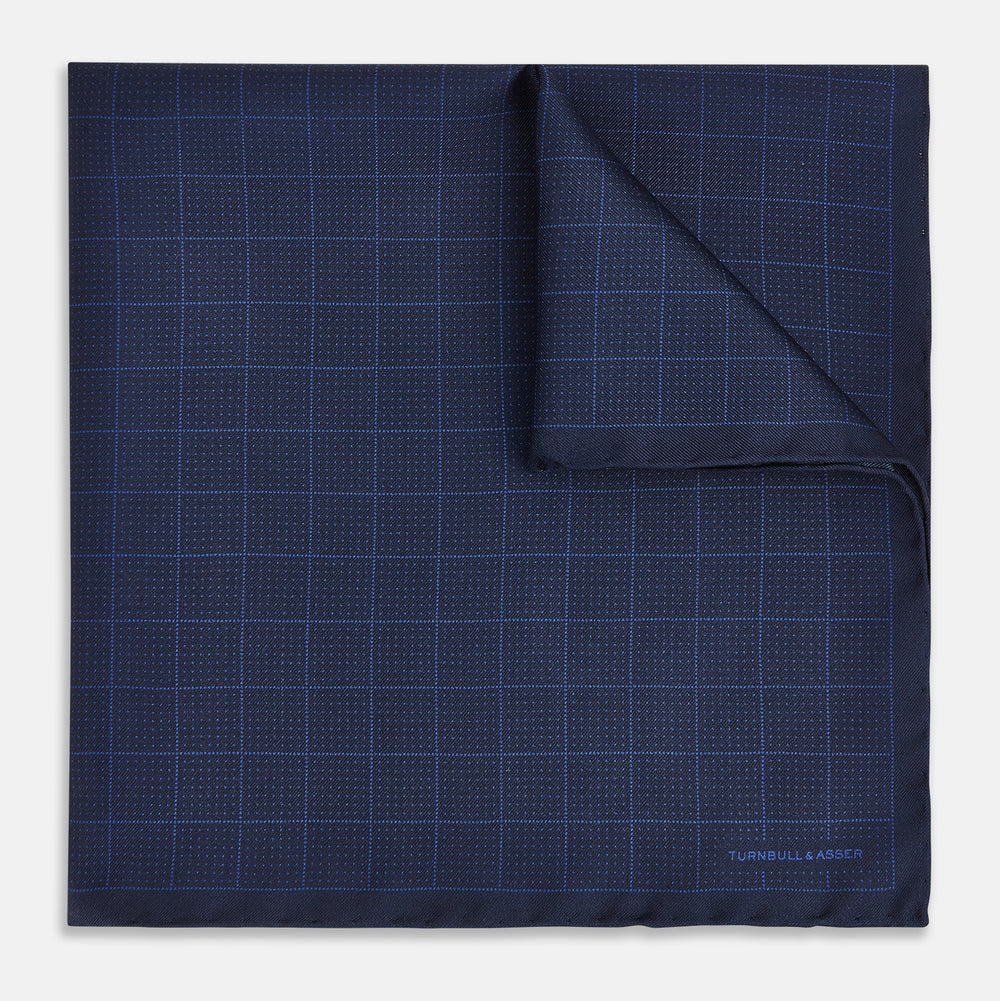 The Checked Blue On Navy Pocket Square in Silk by Turnbull & Asser UK is crafted from dark blue silk and features a subtle geometric design with a lighter blue grid pattern, finished with a discreet Turnbull & Asser logo in the corner.