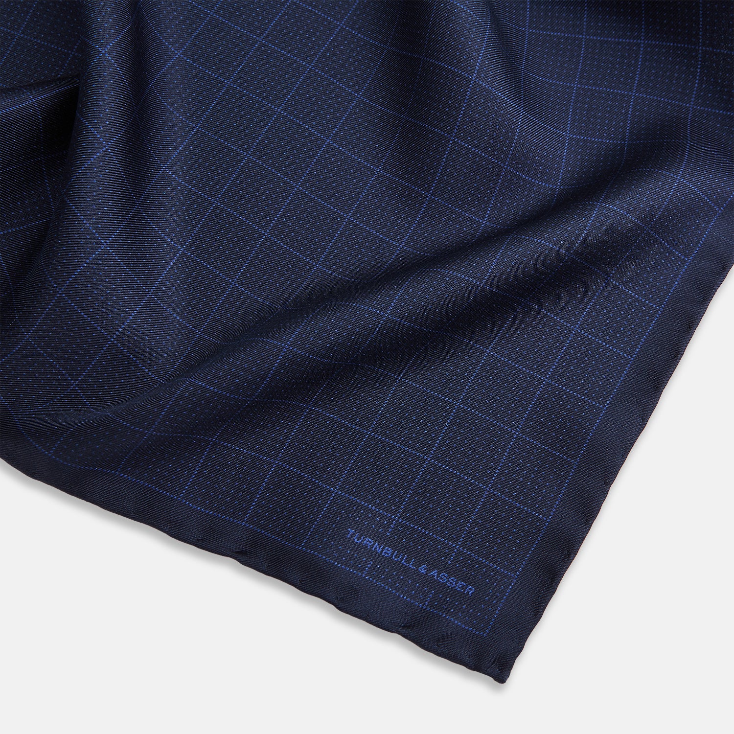 View 2 of product The Checked Blue On Navy Pocket Square in Silk by Turnbull & Asser UK is crafted from dark blue silk and features a subtle geometric design with a lighter blue grid pattern, finished with a discreet Turnbull & Asser logo in the corner.